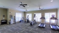 This fitness center offers a thoughtfully designed workout environment spread across a single floor. The space is lined with multiple large windows that allow abundant natural light to fill the room, creating an open and energizing atmosphere for exercise. Three ceiling fans are positioned throughout the ceiling, ensuring consistent airflow and comfort during workouts. Crown molding adds a refined architectural touch to the otherwise functional space.

The room is outfitted with a well-rounded selection of cardio equipment, including treadmills, elliptical trainers, and a stair climber, all arranged to maximize usable floor space. A stationary exercise bike and a stability ball round out the cardio options. Along one wall, a dedicated rack holds a variety of medicine balls, providing options for strength and functional training.

The flooring features durable, patterned carpet tiles suited to a high-traffic fitness environment. Recessed lighting in the ceiling complements the natural light from the windows, ensuring the space remains well-lit at all hours. The neutral, warm-toned walls contribute to a calm and focused workout environment. Overall, this fitness center balances practicality and comfort, making it a functional and inviting space for a variety of exercise routines.