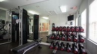 Located at 5700 Magnolia Chase Way in Virginia Beach, Virginia, this single-floor fitness center is thoughtfully equipped to support a wide range of workout routines. The space is divided into distinct functional zones, all unified by durable rubber flooring that provides traction and impact absorption throughout.

The main exercise area features multiple treadmills and elliptical machines, each equipped with individual monitors for an engaging cardio experience. A multi-functional cable machine anchors the strength training section, complemented by a full dumbbell rack ranging across various weights, an adjustable workout bench, and an assortment of medicine balls for dynamic training. Wall-mounted mirrors span much of the space, visually expanding the room while allowing users to monitor their form.

A separate, open flex room is accessible through elegant French doors with louvered shutters, offering a clean, versatile space ideal for stretching, yoga, or mat-based workouts. This area features the same speckled rubber flooring and bright white walls, creating an airy and focused environment.

The facility is well-lit with overhead fluorescent lighting and accented by natural light from windows with slatted blinds. A wall-mounted television in the free weights area adds convenience for entertainment during workouts. The overall design is clean, functional, and professional, making it a well-rounded amenity for residents.
