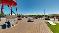 This expansive rooftop deck offers a dynamic outdoor experience defined by its iconic red water tower — a bold architectural landmark bearing the "UTW" insignia that anchors the space visually. The deck is finished with wide-plank composite wood decking throughout, creating a warm, cohesive surface that extends across multiple distinct zones designed for varied uses.

A generous turf lawn area provides a refreshing green contrast to the wood decking and serves as an open gathering or recreation space, complemented by white Adirondack chairs arranged for casual conversation. Nearby, sun loungers with teal cushions offer a dedicated relaxation zone ideal for soaking in the abundant natural light.

Dining and social areas are thoughtfully distributed across the deck, featuring picnic-style tables with bench seating in vibrant orange, as well as bistro-height tables paired with metal chairs — accommodating everything from casual meals to group gatherings. Raised wooden planter boxes introduce greenery throughout, softening the urban aesthetic.

Perimeter metal railings frame the rooftop while preserving unobstructed panoramic views of the surrounding city skyline and treetops. A dedicated restroom facility is conveniently integrated into the rooftop structure. The space benefits from full sun exposure, open sky above, and an energetic urban backdrop, making it a highly functional and visually engaging outdoor venue.