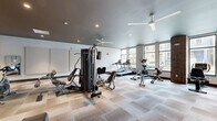 Located at 80 S 18th St. in Columbus, Ohio, this fitness center offers a well-appointed, single-level space designed to support a comprehensive workout experience. The room is finished with durable carpet tile flooring in a neutral, multi-toned pattern that covers the entire area, providing comfort underfoot throughout the space.

The facility is outfitted with a diverse selection of exercise equipment, including treadmills, a multi-station cable weight machine, rowing machines, stationary recumbent bikes, spin cycles, and an elliptical trainer. A wall-mounted flat-screen television is positioned for easy viewing during workouts, and a small open shelving unit provides convenient storage.

Large windows line one side of the room, inviting generous natural light and offering views of the surrounding Columbus streetscape. Exposed brick columns accent the interior, adding a distinctive architectural character that contrasts with the otherwise modern, clean-lined aesthetic. Recessed ceiling lighting and dual ceiling fans ensure the space remains well-lit and well-ventilated at all times.

Full-length mirrors are mounted along one wall, enhancing the sense of openness and allowing users to monitor their form. The overall ambiance is bright, functional, and modern, making this fitness center a practical and inviting amenity for its users.