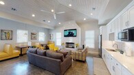 This single-story clubhouse showcases a thoughtfully designed open-concept layout with a distinct coastal character. The main gathering area features soaring vaulted ceilings lined with white-painted shiplap and outfitted with ceiling fans and recessed lighting, creating a bright, airy atmosphere. Polished concrete floors with a marbled finish run throughout, providing a durable yet stylish foundation that ties the space together cohesively.

The living area is anchored by a sleek fireplace set into a dramatic arched white accent wall, above which a large wall-mounted television is prominently displayed. Comfortable seating, including plush gray sofas and bold yellow accent chairs, encourages relaxed gatherings. A casual dining nook features yellow vinyl booth seating arranged near large windows that bring in generous natural light and frame outdoor views.

Coastal-themed artwork, including shorebird paintings and surf-inspired prints, adorns the walls, reinforcing the relaxed seaside ambiance. A well-appointed kitchenette with white shaker cabinetry, tile backsplash, stainless steel appliances, and ample counter space provides convenient amenities for residents and guests. A hallway leads to additional rooms, further expanding the functional offerings of this welcoming community space.
