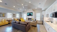 This single-story clubhouse showcases a thoughtfully designed open-concept layout with a distinct coastal character. The main gathering area features soaring vaulted ceilings lined with white-painted shiplap and outfitted with ceiling fans and recessed lighting, creating a bright, airy atmosphere. Polished concrete floors with a marbled finish run throughout, providing a durable yet stylish foundation that ties the space together cohesively.

The living area is anchored by a sleek fireplace set into a dramatic arched white accent wall, above which a large wall-mounted television is prominently displayed. Comfortable seating, including plush gray sofas and bold yellow accent chairs, encourages relaxed gatherings. A casual dining nook features yellow vinyl booth seating arranged near large windows that bring in generous natural light and frame outdoor views.

Coastal-themed artwork, including shorebird paintings and surf-inspired prints, adorns the walls, reinforcing the relaxed seaside ambiance. A well-appointed kitchenette with white shaker cabinetry, tile backsplash, stainless steel appliances, and ample counter space provides convenient amenities for residents and guests. A hallway leads to additional rooms, further expanding the functional offerings of this welcoming community space.