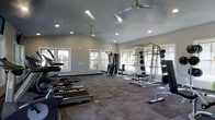 This dedicated fitness center offers a comprehensive workout environment designed to support a wide range of exercise routines. The space features vaulted ceilings that lend an open, airy feel, while ceiling fans ensure comfortable air circulation throughout. Recessed lighting complements the abundant natural light that streams in through multiple windows lining the walls, creating a bright and energizing atmosphere.

The layout is thoughtfully organized to accommodate both cardio and strength training. A row of treadmills and cardio machines lines one side of the room, providing ample space for users to exercise without feeling crowded. On the opposite side, a multi-station cable machine and a full weight rack stocked with plates and barbells cater to strength and resistance training needs. A weight bench is also available for targeted exercises.

The flooring consists of durable, cushioned carpet tiles that offer both comfort underfoot and functional support during workouts. The neutral gray walls and clean, uncluttered design give the space a professional and focused ambiance. Large windows on multiple walls provide views of the surrounding outdoor area, adding a pleasant connection to the outdoors. Overall, this fitness center is a well-rounded facility suited for a variety of fitness goals.
