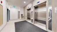 Located at 4118 Cambie Street in Vancouver, BC, this well-appointed building lobby and corridor space offers a clean, contemporary design across a single floor. The entry vestibule welcomes visitors through glass automatic doors, with a runner mat leading into a bright, open foyer finished in large-format white tile throughout. The lobby is equipped with a bank of smart parcel lockers featuring a digital touchscreen interface, alongside a wall-mounted array of individual resident mailboxes framed in light wood paneling — providing efficient mail and package management for building occupants. Two stainless steel elevators, set against warm wood-toned surrounds, offer convenient vertical access, complemented by a nearby water fountain station. The interconnected hallways extend in an L-shaped configuration, with recessed ceiling lighting maintaining a consistently bright and welcoming atmosphere. A split-level transition features low-rise stairs fitted with sleek black metal handrails, adding a refined architectural detail to the circulation path. Floor-to-ceiling glass partitioning along one section of the corridor allows visual openness between zones. Safety signage and security cameras are discreetly integrated throughout. The overall aesthetic is modern and functional, with a neutral white-and-wood palette creating a polished, professional ambiance.
