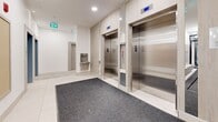 Located at 4118 Cambie Street in Vancouver, BC, this well-appointed building lobby and corridor space offers a clean, contemporary design across a single floor. The entry vestibule welcomes visitors through glass automatic doors, with a runner mat leading into a bright, open foyer finished in large-format white tile throughout. The lobby is equipped with a bank of smart parcel lockers featuring a digital touchscreen interface, alongside a wall-mounted array of individual resident mailboxes framed in light wood paneling — providing efficient mail and package management for building occupants. Two stainless steel elevators, set against warm wood-toned surrounds, offer convenient vertical access, complemented by a nearby water fountain station. The interconnected hallways extend in an L-shaped configuration, with recessed ceiling lighting maintaining a consistently bright and welcoming atmosphere. A split-level transition features low-rise stairs fitted with sleek black metal handrails, adding a refined architectural detail to the circulation path. Floor-to-ceiling glass partitioning along one section of the corridor allows visual openness between zones. Safety signage and security cameras are discreetly integrated throughout. The overall aesthetic is modern and functional, with a neutral white-and-wood palette creating a polished, professional ambiance.