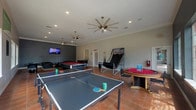 This single-floor game room is a dedicated entertainment and recreation space designed to offer a wide variety of activities for its users. The room is generously sized, with terra cotta tile flooring throughout and crown molding accenting the walls and ceiling. Multiple ceiling fans ensure comfortable air circulation across the entire space.

At the heart of the room sits a full-size pool table with a rich red felt surface, complemented by a nearby cue rack. A ping-pong table occupies one section of the room, offering ample space for competitive play. Additional gaming options include a foosball table, an arcade-style skee-ball or basketball shooting machine, and a dedicated poker table with surrounding bar-height seating.

A lounge area provides a comfortable retreat within the room, furnished with dark leather chairs and a sofa arranged on a striped area rug, all oriented toward a wall-mounted flat-screen television set against a contrasting dark accent wall. Large windows along one side of the room allow abundant natural light to fill the space, offering views of surrounding greenery. A glass block window accent adds an architectural detail to one wall. The overall ambiance is lively and inviting, making this game room a versatile hub for socializing and recreation.