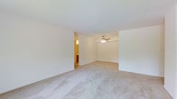 This single-floor space presents a straightforward and functional layout designed for comfortable everyday living. The main living area is open and spacious, featuring wall-to-wall neutral-toned carpeting that lends a warm and inviting feel underfoot. Crisp white walls and a smooth ceiling enhance the sense of brightness throughout the room, while a ceiling fan with an integrated light fixture provides both air circulation and ambient lighting.

The open layout allows for flexible furniture arrangements, making the space adaptable to a variety of needs. A doorway visible from the main area leads into a connected room, which appears to be a bathroom or utility space, featuring warm-toned cabinetry and its own lighting — adding to the overall convenience of the floor plan.

The design is clean and minimal, with no excess ornamentation, allowing the architectural simplicity to stand on its own. Neutral finishes throughout — from the carpeted flooring to the painted walls — create a cohesive and move-in-ready aesthetic. The single-level layout ensures easy accessibility and seamless flow between spaces, making this a practical and well-organized living environment.