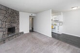 This single-floor space offers a functional and comfortable layout connecting several well-defined areas. The living room serves as a central gathering space, highlighted by a striking floor-to-ceiling stone fireplace anchored in the corner, adding a rustic, textural focal point to the otherwise neutral interior. Soft carpeting covers the living area, contributing to a warm and inviting atmosphere, while dark hardwood-style flooring transitions into the kitchen and dining areas.

The kitchen is designed for efficiency, featuring white cabinetry that extends to the ceiling for ample storage, white appliances including a freestanding electric range and a full-sized refrigerator, and a countertop sink. The layout is galley-style, keeping everything within easy reach. A dining or flex area adjacent to the kitchen benefits from natural light through a window fitted with blinds.

The bathroom is clean and straightforward, equipped with a full tub and shower combination featuring white tile surround, dark wood-style flooring, and a wall-mounted ventilation unit. Textured ceilings are consistent throughout the space, and neutral gray-toned walls provide a cohesive, understated backdrop. The overall design is practical and move-in ready.