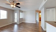 This single-floor residence offers a clean, functional layout centered around a spacious open living area. Rich, dark hardwood-style flooring runs throughout the main living space, providing a warm contrast to the neutral gray walls and bright white baseboards and trim. A ceiling fan with a light fixture is centrally positioned, offering both ventilation and ambient lighting beneath a textured popcorn ceiling.

Natural light fills the room through multiple windows dressed with horizontal blinds, including a glass-paneled exterior door that further brightens the space. The open floor plan connects seamlessly to the kitchen, which features warm wood-tone cabinetry, a counter-height pass-through bar, and a visible range, making it well-suited for both everyday living and entertaining.

A hallway leads toward additional rooms, with glimpses of a lit interior space—likely a kitchen or utility area—visible from the main living room. The layout is practical and well-organized, with clearly defined zones for living, dining, and cooking. Overall, the space presents a straightforward, move-in-ready interior that balances comfort and functionality with classic, understated design choices.