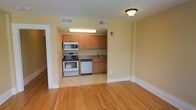 Located at 615 James St in Syracuse, New York, this single-floor space offers a clean, functional layout designed for everyday comfort. The open living area showcases warm hardwood flooring that extends throughout, creating a bright and welcoming atmosphere enhanced by neutral, light-toned walls.

The kitchen is seamlessly integrated into the living space through a defined alcove, providing a natural separation while maintaining an open feel. It is outfitted with stainless steel appliances, including an electric range, built-in microwave, and dishwasher, all complemented by medium-toned wood cabinetry offering ample storage. A single-basin sink sits conveniently beneath overhead cabinets, completing the practical kitchen setup.

Ceiling-mounted light fixtures provide consistent ambient lighting throughout the space, while multiple HVAC vents ensure year-round climate control. Wide baseboards and clean door trim throughout the unit contribute to a classic, well-maintained aesthetic. A fire alarm panel is visible, reflecting attention to safety standards. The overall design balances simplicity and functionality, making the space well-suited for residential living in the heart of Syracuse.