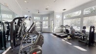 This fitness center is a thoughtfully designed, single-floor facility that offers a comprehensive workout environment for users of all fitness levels. The space features light gray walls accented by crisp white crown molding and baseboards, creating a clean and energizing atmosphere. Large windows with slatted blinds line multiple walls, allowing generous amounts of natural light to fill the room and providing views of lush greenery outside. Ceiling fans contribute to a comfortable climate throughout the space.

The cardio zone is well-stocked with multiple treadmills, elliptical trainers, and a stationary bike, all positioned to take advantage of the natural light and outdoor views. Wall-mounted televisions provide entertainment during workouts. The strength training area features a multi-station cable machine, a dedicated free weights section with a full rack of dumbbells in varying sizes, and additional resistance machines targeting different muscle groups. Full-length mirrors along one wall visually expand the space and allow users to monitor their form.

The durable rubberized flooring throughout provides cushioning and safety underfoot. The layout is spacious and well-organized, ensuring ease of movement between equipment. With its bright, airy design and diverse equipment offerings, this fitness center delivers a professional and motivating environment for daily exercise.