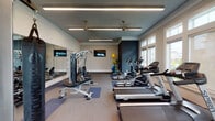Located at 3900–3940 Cimarron Blvd in Corpus Christi, Texas, this single-story fitness center is thoughtfully designed to support a comprehensive workout experience. The space features a tray ceiling with a teal-painted recess, ceiling fans, and linear LED lighting that brightens the room evenly throughout the day.

Natural light is a defining characteristic of this facility, provided by a generous bank of large, multi-pane windows along one wall that offer outdoor views and create an open, airy atmosphere. The flooring consists of a combination of carpet and modular fitness mats in blue and gray tones, delineating different workout zones.

Cardio equipment includes multiple treadmills, elliptical trainers, and a stationary bike, all positioned to take advantage of the natural light from the windows. A rowing machine is situated in a quieter corner alongside framed artwork, adding a refined aesthetic touch. The strength training area features a multi-station cable machine, a weight bench, dumbbell racks with a full range of free weights, and a heavy punching bag with a ceiling-mounted bracket. A large wall mirror expands the visual depth of the space. A medicine ball rack with colorful balls adds a functional and vibrant accent. White built-in shelving provides storage, contributing to the clean, organized layout.