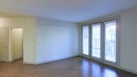 This single-floor space is designed with a clean, modern aesthetic that emphasizes openness and natural light. The main living area features rich, dark hardwood-style flooring that contrasts attractively with the soft, neutral-toned walls, creating a warm yet contemporary atmosphere throughout.

A prominent feature of the space is the set of full-length glass French doors positioned along one wall. Fitted with horizontal blinds for privacy and light control, these doors open to an outdoor balcony, allowing abundant natural light to pour into the interior while offering a seamless connection between indoor and outdoor living.

The walls are finished in a light, neutral palette, providing a versatile backdrop suitable for a variety of interior styles. White baseboards frame the perimeter of the room, adding a crisp, finished look. Multiple electrical outlets are positioned conveniently along the walls, reflecting thoughtful layout planning.

The ceiling is smooth and bright, further enhancing the sense of airiness within the space. Overall, the design balances simplicity and functionality, making it an adaptable, light-filled environment well-suited to modern living.