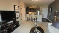 This single-floor residence features an open-concept layout that connects the living area and kitchen into one cohesive, functional space. The living area is anchored by a dark-finished media console and adorned with framed artwork, adding a refined aesthetic to the neutral-toned walls and plush carpeted flooring. A round accent table with decorative sculptural pieces in the foreground adds an elegant finishing touch.

The kitchen is a standout feature, showcasing dark espresso cabinetry that contrasts beautifully with light granite countertops. A peninsula with a raised breakfast bar accommodates two cream-upholstered bar stools, serving as both a casual dining option and a natural divider between the cooking and living zones. Stainless steel appliances, including a refrigerator and microwave, add a modern, polished element. Track lighting overhead ensures the kitchen is well-illuminated, enhancing both functionality and ambiance.

A hallway visible in the background hints at additional rooms beyond, while a secondary door provides convenient access. The overall palette of warm neutrals, dark wood tones, and soft textiles creates a sophisticated yet welcoming atmosphere throughout.