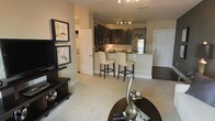 This single-floor residence features an open-concept layout that connects the living area and kitchen into one cohesive, functional space. The living area is anchored by a dark-finished media console and adorned with framed artwork, adding a refined aesthetic to the neutral-toned walls and plush carpeted flooring. A round accent table with decorative sculptural pieces in the foreground adds an elegant finishing touch.

The kitchen is a standout feature, showcasing dark espresso cabinetry that contrasts beautifully with light granite countertops. A peninsula with a raised breakfast bar accommodates two cream-upholstered bar stools, serving as both a casual dining option and a natural divider between the cooking and living zones. Stainless steel appliances, including a refrigerator and microwave, add a modern, polished element. Track lighting overhead ensures the kitchen is well-illuminated, enhancing both functionality and ambiance.

A hallway visible in the background hints at additional rooms beyond, while a secondary door provides convenient access. The overall palette of warm neutrals, dark wood tones, and soft textiles creates a sophisticated yet welcoming atmosphere throughout.