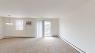 This single-floor unit offers a straightforward, functional layout across several well-defined spaces. The living area is generously proportioned, featuring neutral wall-to-wall carpeting, crisp white walls, and a sliding glass door fitted with vertical blinds that invites natural light and provides access to an outdoor area. A wall-mounted air conditioning unit and baseboard heating ensure year-round climate control throughout the carpeted rooms.

The kitchen is separated from the living area by a partial wall, creating a semi-open feel. It features white cabinetry with ample upper and lower storage, light countertops, a stainless steel sink, and white appliances including a gas range with oven and a full-size refrigerator. Hardwood-style flooring completes the kitchen space, offering a practical contrast to the carpeted areas.

The bedroom is a quiet, well-lit room with its own wall-mounted A/C unit, baseboard heating, and a window with blinds. The bathroom features a full-size soaking tub with tiled surround, and a clean, white aesthetic throughout. The overall ambiance of the unit is neutral and move-in ready, providing a clean canvas for personalization.