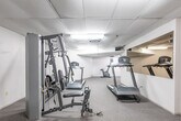 Located at 1850 Rathburn Rd E in Mississauga, Ontario, this fitness facility offers a well-organized exercise space designed to accommodate a range of workout needs. The room features a drop-tile ceiling with recessed fluorescent lighting that provides consistent, even illumination throughout. The flooring is finished with durable, low-pile carpet suitable for fitness use.

The cardio zone is the focal point of the space, housing multiple treadmills and upright stationary bikes arranged to maximize use of the floor area. A wall-mounted mirror panel runs along one side, creating a sense of depth and allowing users to monitor their form during exercise. The strength training area features a multi-function cable and weight machine, offering a variety of resistance-based exercises within a compact footprint.

A small window on one wall provides a modest source of natural light, complementing the overhead fixtures. The neutral white walls keep the environment clean and functional. Access to the facility is provided through a standard door with emergency signage, ensuring safety compliance. Overall, the space is efficiently laid out to deliver a practical and accessible fitness experience for residents of the building.