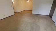 This single-floor space presents a straightforward, open layout centered around a carpeted living area. The flooring throughout is wall-to-wall carpet in a neutral beige tone, providing a soft underfoot feel across the generous open floor plan. The walls are painted in a light, neutral shade, contributing to a clean and understated aesthetic that serves as a blank canvas for a variety of interior styles.

A ceiling fan is centrally positioned overhead, offering both air circulation and a subtle decorative element to the room. Dark baseboards provide a mild contrast against the lighter walls, adding a modest defining detail to the overall interior. A wall-mounted light fixture is visible along one side, supplementing the room's lighting options.

The layout features an L-shaped or segmented wall configuration, creating a natural division within the open space that could accommodate distinct functional zones. Electrical outlets are conveniently placed along the walls. The space is currently vacant, making it easy to envision custom furnishing arrangements. Overall, the room offers a practical, move-in-ready environment with classic, neutral finishes throughout.