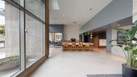 This contemporary lobby presents a striking first impression through its expansive floor-to-ceiling glass facade, which floods the interior with natural light and offers open views of the surrounding streetscape. The polished terrazzo flooring reflects this abundant light, enhancing the sense of openness throughout the space. A central communal table with warm-toned leather chairs serves as a functional gathering point, complemented by a built-in credenza featuring wood cabinetry and lush potted plants that add a natural element to the design. A textured chevron-patterned accent wall provides a sophisticated focal point, while large mirror panels amplify depth and brightness. Exposed concrete wall elements contrast with the refined interior finishes, lending an industrial character that balances the lobby's modern aesthetic. A dedicated mailroom area with organized built-in cubbies and a warm wood surround is seamlessly integrated into the layout. An adjacent glass-enclosed meeting or office room is visible, offering flexible workspace. Decorative touches, including a circular wooden wall art piece, contribute to the curated, design-forward atmosphere. The overall ambiance is clean, open, and welcoming, combining functional amenities with thoughtful architectural detailing.