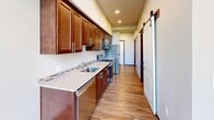 This efficiently designed single-floor unit makes smart use of its compact footprint across one bedroom, one bathroom, two kitchen areas, a closet, and connecting hallways. Light wood-look flooring runs consistently throughout, lending visual continuity and warmth to the space.

The kitchen is a standout feature, designed in a galley configuration with rich, dark wood cabinetry extending to the ceiling, light quartz-style countertops with a tile backsplash, stainless steel appliances including a range, dishwasher, and refrigerator, and a single-basin sink. A sliding barn door adds a modern, space-conscious design touch at the kitchen's entrance.

The bedroom, though compact, benefits from impressively tall ceilings and large windows that introduce abundant natural light and open views of surrounding greenery, creating a sense of spaciousness beyond its physical dimensions.

The bathroom is well-appointed with a single-sink vanity featuring warm wood cabinetry and a light stone-style countertop, a walk-in shower with grab bars, and a stacked washer and dryer conveniently integrated into the space—eliminating the need for a separate laundry area. Neutral wall tones throughout maintain a clean, modern aesthetic.