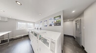 This single-floor shared laundry facility is designed with functionality and cleanliness in mind. The space features a bright, modern aesthetic defined by crisp white walls, recessed ceiling lighting, and durable dark slate-toned tile flooring throughout. The main laundry area is well-equipped with a row of Coinamatic-branded Huebsch front-load washers and top-load dryers, offering ample capacity for residents. Each machine is clearly numbered and accompanied by posted operating instructions, making the space straightforward to use. A built-in folding counter along one wall provides a convenient surface for sorting and folding laundry. Natural light filters in through windows, contributing to the room's open and airy feel. Informational signage is displayed throughout, including details about the Coinamatic smart laundry portal, which allows users to manage laundry payments and machine availability digitally. A secure, keypad-controlled entry door ensures controlled access to the facility. A connecting corridor with recessed lighting and matching tile flooring links the laundry room to the broader building. The overall ambiance is clean, organized, and utilitarian, providing a practical and comfortable shared amenity space.
