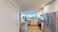 This contemporary single-floor residence offers a thoughtfully designed open-concept layout that maximizes its compact footprint. Light wood-tone flooring flows consistently throughout, enhancing the cohesive and airy aesthetic across all rooms.

The kitchen is outfitted with sleek flat-panel white cabinetry, stainless steel appliances including a gas range and French door refrigerator, a white subway tile backsplash, and a stone-topped island with seating — ideal for both casual dining and meal preparation. The kitchen opens directly into the living area, which is bright and spacious, featuring floor-to-ceiling sliding glass doors that lead to a private balcony with outdoor views of the surrounding cityscape and greenery.

The bedroom is a calm retreat with large windows on two walls, allowing ample natural light and tree-lined views. It comfortably accommodates a full sleeping and workspace setup. The bathroom features a clean, minimalist design with a stone-topped vanity, integrated sink, large mirror, and wood-toned cabinetry.

The entryway is clean and functional, with in-unit storage access nearby. Recessed lighting throughout the unit contributes to a bright, well-lit interior. The overall ambiance is modern and refined, combining clean architectural lines with a warm, livable atmosphere.