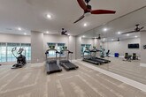 Located at 3904 Durham Park Rd in Stone Mountain, Georgia, this fitness center offers a thoughtfully designed workout space within a compact, single-floor footprint. The interior is finished with neutral gray walls, white baseboards, and durable patterned carpet tiles that lend a clean, modern aesthetic throughout.

The main exercise area is anchored by multiple treadmills and an elliptical trainer, positioned along a wall of large windows that invite natural light and views of the surrounding greenery. A full-length wall of floor-to-ceiling mirrors visually expands the space while allowing users to monitor their form during workouts. Two ceiling fans with dark wood-tone blades are mounted overhead, providing consistent air circulation.

A dedicated free weights and strength training zone includes a weight bench, color-coded medicine balls, and a cable resistance machine, all neatly organized. A wall-mounted flat-screen television provides entertainment during workouts. A stationary bike rounds out the cardio offerings.

Near the entrance, a posted fitness center rules board and a hand sanitizer dispenser reflect attention to cleanliness and safety. The entry area also features a rack of medicine balls and leads to an adjacent corridor. Recessed lighting throughout ensures the space is consistently well-lit, creating a functional and welcoming environment for residents to maintain an active lifestyle.