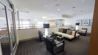 This single-floor space presents a polished and professional interior characterized by clean lines and a contemporary design aesthetic. The lounge area serves as a central hub, thoughtfully furnished with upholstered armchairs and sofas in neutral tones of cream and charcoal, complemented by dark-framed coffee and side tables. A bold floor lamp with a cylindrical shade anchors the seating arrangement, adding both style and ambient lighting to the space.

The interior benefits from an abundance of natural light streaming through large windows fitted with white roller blinds, creating a bright and airy atmosphere throughout. White ceilings with recessed lighting further enhance the sense of openness, while structural columns are seamlessly integrated into the design.

A half-wall partition with a timber rail separates the lounge from an adjacent reception or service counter area, which features a clean, minimal desk setup and additional windows overlooking greenery outside. The flooring transitions between low-pile patterned carpet in the lounge zone and what appears to be a smooth surface near the service area, adding visual definition to the different functional zones.

Subtle decorative touches, including potted plants and framed artwork, lend warmth and personality to an otherwise crisp, modern environment. Overall, the space conveys a professional yet inviting ambiance suited to hospitality or corporate use.