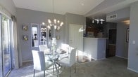 This single-floor space showcases an open-concept layout that seamlessly connects the dining area and kitchen. The dining area is anchored by a round glass-top table with chrome legs, surrounded by sleek white chairs, and illuminated by a striking multi-bulb sputnik-style chandelier that serves as a modern centerpiece. Grey walls throughout create a cohesive, neutral palette that complements the contemporary aesthetic.

Natural light filters in through a glass door and sidelight panel, brightening the carpeted dining space and highlighting the table setting. The kitchen, partially separated by a half-wall breakfast bar with a light countertop, features warm wood cabinetry, a microwave, and track lighting overhead. Dark hardwood flooring in the kitchen contrasts with the carpeted dining area, subtly defining each zone.

The overall ambiance is modern and understated, with clean lines and a neutral color scheme that provides a versatile foundation. The open sightlines between the kitchen and dining area enhance the sense of space, making it well-suited for both everyday living and casual entertaining.
