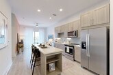 This thoughtfully laid-out one-bedroom, one-bathroom residence offers an efficient and stylish single-floor plan with a cohesive modern aesthetic throughout. The open-concept kitchen and living area form the heart of the space, where light wood-toned cabinetry, a subway tile backsplash, quartz countertops, and a full suite of stainless steel appliances create a functional yet refined cooking environment. A central kitchen island with built-in shelving doubles as a breakfast bar, providing additional seating and storage while seamlessly connecting to the living area. Light wood-style plank flooring flows throughout the main living spaces, complemented by recessed lighting and a ceiling fan that enhance the bright, airy atmosphere. A large window at the end of the living area allows natural light to pour in, adding to the open feel of the space. The bedroom features plush carpet, a warm accent wall, and a window with sheer curtains that invite soft, natural light. The full bathroom is appointed with a wood-toned vanity, quartz countertop, and a soaking tub with tile surround, and connects directly to a spacious walk-in closet with built-in shelving and hanging rods. A dedicated laundry area completes this well-equipped residence.