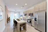 This thoughtfully laid-out one-bedroom, one-bathroom residence offers an efficient and stylish single-floor plan with a cohesive modern aesthetic throughout. The open-concept kitchen and living area form the heart of the space, where light wood-toned cabinetry, a subway tile backsplash, quartz countertops, and a full suite of stainless steel appliances create a functional yet refined cooking environment. A central kitchen island with built-in shelving doubles as a breakfast bar, providing additional seating and storage while seamlessly connecting to the living area. Light wood-style plank flooring flows throughout the main living spaces, complemented by recessed lighting and a ceiling fan that enhance the bright, airy atmosphere. A large window at the end of the living area allows natural light to pour in, adding to the open feel of the space. The bedroom features plush carpet, a warm accent wall, and a window with sheer curtains that invite soft, natural light. The full bathroom is appointed with a wood-toned vanity, quartz countertop, and a soaking tub with tile surround, and connects directly to a spacious walk-in closet with built-in shelving and hanging rods. A dedicated laundry area completes this well-equipped residence.