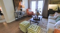 This single-floor space offers a warm and inviting atmosphere centered around a thoughtfully designed living area. Rich hardwood flooring runs throughout, grounding the space with a natural, earthy tone that complements the overall aesthetic. Large windows dressed with patterned curtains flood the room with natural light, creating a bright and airy ambiance.

The living area is arranged for both comfort and visual interest. A dark wood coffee table serves as the focal point, styled with a decorative tray, candles, and a floral arrangement that adds a touch of elegance. Two bold green-and-white geometric floor poufs provide flexible, casual seating and introduce a lively pop of color. An accent chair in a warm terracotta hue adds a cozy corner nook beside the windows, complemented by a striped throw pillow.

Along one wall, a sleek industrial-style open shelving unit displays curated décor items, including vases, framed photos, and a sculptural lamp, adding personality and depth to the room. A striking wall art piece featuring a bamboo motif anchors the space with a dramatic, nature-inspired focal point. The overall design balances eclectic accents with cohesive, neutral tones, resulting in a space that feels both curated and livable.