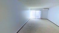 This single-floor space presents a clean, open layout designed for versatility and practicality. The main area is finished with wall-to-wall carpeting in a neutral beige tone, providing a soft and understated foundation throughout. White walls keep the interior bright and adaptable, while a drop-panel ceiling adds a classic, functional overhead finish.

A prominent feature of the space is the large sliding glass window dressed with vertical blinds, which serves as the primary source of natural light. When open, the blinds allow generous daylight to flood the interior, creating a welcoming and airy atmosphere. The window also provides an outward view, connecting the interior to the exterior surroundings.

A wall-mounted air conditioning unit is positioned near the window, ensuring climate control comfort throughout the space. The overall layout is unobstructed and open, making the area highly adaptable to a variety of uses, whether as a living area, workspace, or multipurpose room. The combination of neutral finishes, natural light, and practical amenities gives this space a straightforward, functional character well-suited to everyday use.