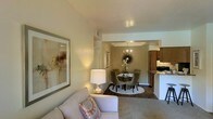 This single-floor residence offers a thoughtfully designed open-concept layout that seamlessly connects the main living areas. The living room is a welcoming space featuring neutral-toned carpet, smooth walls with a subtle warm finish, and a ceiling-mounted ventilation vent. Décor-ready wall space is complemented by soft, ambient lighting, creating a relaxed and inviting atmosphere.

The living area flows naturally into the dining space, which is defined by a tray ceiling detail and an elegant chandelier overhead. The dining area features hard surface flooring and provides an intimate setting for meals and gatherings. A round dining table with upholstered chairs anchors the space, positioned beneath tasteful wall décor.

Adjacent to the dining area is the kitchen, which is accessible via a counter-height bar-style opening, allowing for easy interaction between spaces. The kitchen features warm-toned cabinetry and functional counter space, making it practical for everyday use.

A hallway connects the living spaces to the private quarters, leading to a bedroom with warm lighting and an adjoining bathroom. Throughout the residence, the neutral color palette, consistent finishes, and cohesive design choices create a calm, unified ambiance suited for comfortable everyday living.
