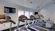 Located at 2000 Varsity Lane in Bear, Delaware, this single-floor fitness center is a well-appointed exercise facility designed to support an active lifestyle. The space is equipped with a row of treadmills and elliptical machines, providing residents with a variety of cardio workout options. Multiple wall-mounted flat-screen televisions are positioned throughout the room, allowing users to stay entertained during their workouts. Natural light filters in through a series of windows fitted with horizontal blinds, creating a bright and energizing atmosphere. Recessed ceiling lighting supplements the natural light, ensuring the space remains well-lit at all times. The flooring features a striped, multi-toned carpet that adds a modern touch to the interior. The walls are finished in a clean, neutral white, contributing to the open and airy feel of the room. The vaulted ceiling adds a sense of spaciousness to the facility. Overall, the fitness center offers a functional and comfortable environment well-suited for daily exercise routines.