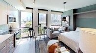Located at 33 N St NE in Washington, DC's vibrant NoMa neighborhood, this modern studio is thoughtfully designed to maximize both function and style. The open-plan living area is anchored by a wall of large, floor-to-ceiling windows that flood the space with natural light and frame sweeping views of the surrounding cityscape, including neighboring green tree canopies and urban skyline. Light hardwood-style flooring runs throughout, complementing a contemporary aesthetic with cool teal accent walls and clean white finishes.

The sleeping area comfortably accommodates a full bed and includes a dedicated workspace with a desk and chair, as well as wall-mounted media storage — ideal for working from home. A high-top bistro table near the windows creates a secondary dining or casual work zone. The kitchen is efficiently laid out with wood-grain lower cabinetry, white upper cabinets, light quartz countertops, subway tile backsplash, a stainless steel sink, and full appliances including an electric range, built-in microwave, and refrigerator. The bathroom features large-format tile flooring, a glass-enclosed shower, a floating wood-accented vanity with an undermount sink, and clean, neutral finishes throughout.