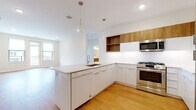 This single-floor residence showcases a thoughtfully designed open-concept layout that seamlessly connects the kitchen and living area. The kitchen serves as the focal point, featuring a large peninsula with a crisp white quartz countertop and an integrated sink. Cabinetry combines flat-front white lower units with warm wood-toned upper cabinets and open shelving, creating a contemporary two-tone aesthetic. Stainless steel appliances include a gas range, over-range microwave, French door refrigerator, and dishwasher. Light hardwood flooring runs throughout the main living area, complementing the recessed lighting and pendant fixtures overhead. Natural light filters in through windows that overlook the surrounding neighborhood.

The bedroom is finished with plush carpet and features dual windows that provide ample natural light. An adjacent walk-in closet offers wire shelving and hanging rods for generous storage. The bathroom is clean and modern, with light-toned large-format tile flooring, a wood-front vanity with a white countertop, and a well-lit mirror. A dedicated in-unit laundry area accommodates a full-sized Whirlpool front-loading washer and dryer, adding practical convenience to this well-appointed space.