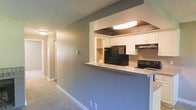 This single-floor space showcases a practical and open layout that connects the kitchen and living areas in a cohesive flow. The kitchen is defined by a raised peninsula counter with a light-toned surface, serving as both a functional workspace and a natural divider between the cooking and living zones. White upper cabinetry lines the kitchen walls, providing ample storage, while black appliances — including a refrigerator and range with an overhead ventilation hood — add contrast to the lighter surroundings. A dedicated overhead light fixture illuminates the kitchen area, set within a recessed ceiling detail that adds subtle architectural interest. The interior walls feature a textured finish in a neutral tone, contributing to a warm and understated ambiance throughout. Carpeted flooring extends through the living area, adding comfort underfoot. A hallway visible from the main space leads further into the unit, suggesting additional rooms beyond. The overall design emphasizes efficient use of space, with a straightforward and functional aesthetic that is well-suited for everyday living.