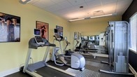 This fitness center offers a clean, functional environment tailored for cardiovascular and general exercise. The space is finished with soft yellow walls and a drop ceiling with recessed fluorescent lighting that ensures consistent, bright illumination throughout. The flooring features a durable, low-pile carpet with striped detailing, providing traction and comfort during workouts.

The room is outfitted with several Precor cardio machines, including treadmills and an elliptical trainer, each equipped with individual digital display monitors for tracking workout metrics. Framed artwork adorns the walls, adding a modest decorative touch to the otherwise utilitarian space.

A full wall of mirrors along one side of the room visually expands the space and allows users to monitor their form during exercise. The mirrors also reflect the equipment, giving the gym a more spacious feel. Ventilation is provided through ceiling vents, supporting air circulation during active use.

Overall, the fitness center presents a straightforward, practical layout that prioritizes usability, making it a convenient and accessible option for those seeking a dedicated space for daily cardio and exercise routines.