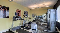 This fitness center offers a clean, functional environment tailored for cardiovascular and general exercise. The space is finished with soft yellow walls and a drop ceiling with recessed fluorescent lighting that ensures consistent, bright illumination throughout. The flooring features a durable, low-pile carpet with striped detailing, providing traction and comfort during workouts.

The room is outfitted with several Precor cardio machines, including treadmills and an elliptical trainer, each equipped with individual digital display monitors for tracking workout metrics. Framed artwork adorns the walls, adding a modest decorative touch to the otherwise utilitarian space.

A full wall of mirrors along one side of the room visually expands the space and allows users to monitor their form during exercise. The mirrors also reflect the equipment, giving the gym a more spacious feel. Ventilation is provided through ceiling vents, supporting air circulation during active use.

Overall, the fitness center presents a straightforward, practical layout that prioritizes usability, making it a convenient and accessible option for those seeking a dedicated space for daily cardio and exercise routines.