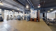 Located at 310 Granello Ave in Coral Gables, Florida, this expansive single-floor fitness center offers a comprehensive exercise environment designed to accommodate a wide range of workout needs. The space features a modern industrial aesthetic, with exposed ductwork running across dark-painted ceilings, pendant lighting, and structural columns that define the open floor plan.

The cardio zone is prominently positioned near a striking wall of floor-to-ceiling windows, flooding the area with natural light and offering views of the surrounding neighborhood. This section is outfitted with a robust selection of Matrix-brand treadmills, ellipticals, stair climbers, and stationary cycling bikes arranged in organized rows.

The strength-training area occupies the interior of the facility and includes a full array of selectorized machines, cable systems, free-weight benches, barbells, and a dumbbell rack. Large wall-mounted mirrors extend across multiple sections, enhancing the sense of space and allowing users to monitor their form.

A dedicated free weights and functional training zone features rubber flooring for impact absorption, alongside storage lockers for personal belongings. Wall-mounted televisions are positioned throughout the facility. The neutral-toned flooring, white walls, and abundant equipment create a professional, gym-quality environment suited for serious fitness training.