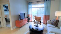 This single-floor living space is thoughtfully designed with a contemporary aesthetic and a warm, vibrant color palette. The living area centers around a glossy coral-orange media console, which houses a flat-screen television and serves as a bold focal point against the soft grey walls. Decorative candlestick holders flank the television, adding an elegant touch.

Natural light fills the room through a large sliding glass door fitted with sheer light-blue curtains accented by coral-orange panels, which complement the room's overall color scheme. The door opens to a balcony with a railing, offering a pleasant outdoor view of surrounding greenery and trees, enhancing the sense of openness within the space.

The living area accommodates a round dark-wood coffee table, styled with decorative books and ornamental floral arrangements, positioned atop a patterned area rug over light hardwood-style flooring. A neutral-toned armchair with decorative spherical accents completes the seating arrangement. A potted plant near the balcony door adds a natural, refreshing element to the interior.