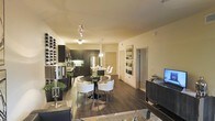 This single-floor residence showcases a thoughtfully designed open-concept layout that seamlessly connects the kitchen, dining, and living areas into one cohesive and inviting space. Rich hardwood flooring runs throughout, anchoring the contemporary aesthetic and lending warmth to the interior.

The modern kitchen is outfitted with sleek dark cabinetry, stainless steel appliances, and a kitchen island with pendant lighting, creating both a functional and visually striking culinary space. A built-in wine rack along the kitchen wall adds a refined, lifestyle-oriented touch for entertaining enthusiasts.

Adjacent to the kitchen, the dining area features a round glass-top table paired with elegant white chairs, illuminated by a chic overhead light fixture. The space flows naturally into the living area, where a media console and comfortable seating complete the arrangement.

Track lighting along the ceiling provides flexible, ambient illumination throughout the main living space, while neutral-toned walls keep the atmosphere bright and airy. A hallway leads to additional rooms beyond, suggesting a well-organized floor plan. The overall ambiance is modern and sophisticated, with a careful balance of dark and light design elements that create a polished, upscale feel throughout the home.