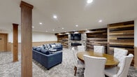 Located at 1960 PA-611 in Swiftwater, Pennsylvania, this single-floor game room offers a well-appointed communal space designed for relaxation and social activity. The room's defining feature is a striking accent wall composed of reclaimed wood planks in varied earth tones, anchored by a stacked-stone fireplace with a mounted flat-screen television above — creating a warm, lodge-inspired focal point. The space is furnished with two navy blue sofas adorned with patterned throw pillows, arranged to face the fireplace for an inviting lounge area ideal for conversation or entertainment. A round wooden dining table surrounded by upholstered chairs provides a dedicated spot for games or casual dining, complemented by a vintage-style "Game Room" sign that reinforces the space's recreational character. Structural wooden columns add architectural interest throughout the room, while a two-tone wall treatment — warm tan wainscoting below and crisp white above — contributes to the cohesive, welcoming aesthetic. Recessed lighting illuminates the room evenly across the patterned wall-to-wall carpeting. Additional features include access to a hallway with supplementary doors and storage areas. The overall ambiance is casual yet thoughtfully designed, blending rustic and contemporary elements to create a comfortable, multi-purpose gathering space.