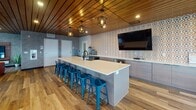 Located at 1001 Belknap St in Superior, Wisconsin, this single-floor communal lounge space combines modern design with warm, inviting materials throughout. The interior is unified by rich wood-plank ceilings that run the length of the space, complemented by durable hardwood-style flooring underfoot.

The kitchen and bar area serves as a central hub, featuring a large island topped with white quartz and lined with vibrant blue metal barstools. A bold geometric-patterned accent wall in warm tones anchors the space behind the island, while geometric pendant lights hang overhead. Gray cabinetry, a built-in sink, and a wall-mounted TV complete this functional yet stylish gathering area.

The lounge zone is generously proportioned, centered around a substantial burnt-orange leather sectional sofa with natural wood stump side tables. A striking circular halo light fixture above adds a dramatic focal point. Large floor-to-ceiling windows and glass doors flood the space with natural light and open onto an outdoor terrace, offering sweeping views of the surrounding Superior skyline.

The game and recreation area includes a pool table and shuffleboard, making it ideal for social gatherings. A wall-mounted TV with a floating wood media console provides an additional entertainment option. Throughout, the design balances industrial and natural elements, creating a warm, modern atmosphere suited for community interaction.