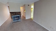 This single-floor residence offers a straightforward and functional layout designed for comfortable everyday living. The living area serves as the heart of the space, highlighted by a striking stone-surround fireplace with a glass-front insert, adding a classic, rustic focal point to the room. Neutral-toned wall-to-wall carpeting runs throughout, contributing to a warm and cohesive atmosphere. The textured ceiling adds a layer of traditional character consistent throughout the space. A central hallway connects the main living area to the rest of the home, providing easy circulation between rooms. Adjacent to the living area, the kitchen is visible through the open layout and features light wood cabinetry, white appliances, and tile flooring, offering a practical and well-organized cooking space. A ventilation register is positioned high on the wall, indicating a central HVAC system for year-round climate control. The neutral, light beige color palette across the walls and flooring creates a clean, move-in-ready canvas. Overall, the space presents a simple yet inviting single-level layout suited for a variety of living arrangements.