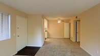 This single-floor space features an open-concept layout that connects the main living area to the kitchen and additional rooms. The living area is finished with neutral-toned wall-to-wall carpeting, contributing to a warm and understated ambiance. Smooth, light-colored walls throughout the space provide a clean backdrop, while white trim along the baseboards and door frames adds a crisp, finished look.

A ceiling fan is centrally positioned overhead, providing both lighting and air circulation to the main area. The entryway transitions from dark hardwood-style flooring into the carpeted living space, creating a subtle delineation between zones.

Visible from the main living area, the kitchen features white cabinetry and appears compact yet functional, with a sink and counter space noted along the far wall. Multiple doorways lead to additional rooms, suggesting a practical floor plan with accessible privacy. Vents are visible along the walls, indicating a ducted HVAC system. Overall, the space presents a straightforward, move-in-ready layout suited for comfortable everyday living.