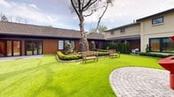 Located at 1188 King St in Greenwich, Connecticut, this single-story property showcases a thoughtfully designed architectural composition that seamlessly blends contemporary materials with natural surroundings. The exterior is defined by warm vertical wood cladding paired with crisp dark trim and rooflines, creating a modern yet grounded aesthetic that complements the lush landscape.

At the heart of the property lies a central courtyard, which serves as a defining outdoor feature. Lush, manicured green lawn extends across the courtyard, anchored by a circular stone paver installation at its center, where a vibrant red abstract sculpture commands immediate attention. Mature trees rise above the roofline, providing a natural canopy and a sense of established character. Wooden garden benches are positioned throughout the space, offering comfortable areas to take in the surroundings.

The building's wings wrap around the courtyard on multiple sides, with large multi-pane windows framed in dark metal providing generous views into and out of the interior spaces. Folding glass doors on one wing open directly onto a stone-paved terrace, facilitating a fluid connection between indoor and outdoor living. Neatly maintained garden beds featuring evergreen shrubs and ornamental plantings line the building's perimeter, adding further greenery and texture to the overall design.