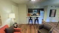 This single-floor residence offers an open-concept layout that seamlessly connects the living and kitchen areas, creating a cohesive and functional environment. The kitchen is a standout feature, equipped with stainless steel appliances including a gas range, built-in microwave, and a full-size refrigerator. Dark espresso-toned cabinetry provides a striking contrast against the light countertops, while a peninsula with bar seating for two serves as both a prep space and a casual dining area. Track lighting above the kitchen ensures ample task illumination. The flooring throughout is a warm hardwood-style material, adding visual continuity and a sense of warmth to the space. The living area accommodates comfortable seating and is complemented by a ceiling fan for climate control. Neutral green-toned walls provide a calm backdrop, while recessed and pendant lighting enhance the overall ambiance. The layout is practical and well-organized, making efficient use of the available space while maintaining a welcoming and modern aesthetic throughout.
