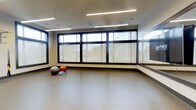 The fitness studio at Station Village at Avenel is a well-appointed, single-floor exercise space designed to support a variety of workout routines and wellness activities. The room is bright and open, benefiting from a generous bank of large, dark-framed windows along the exterior wall that allow abundant natural light to fill the space while offering views to the outdoors. Roller shades provide residents with the option to control light and privacy as needed. The studio features smooth, resilient flooring ideal for yoga, stretching, and dynamic movement exercises. A full-length mirror spans one wall, a practical and functional element for form-checking during workouts. The ceiling is clean and contemporary, lined with recessed lighting fixtures and integrated ceiling speakers, ensuring the space is well-lit and conducive to energetic exercise sessions at any time of day. Exercise equipment such as stability balls is available within the room, reflecting the studio's versatility as a multi-use fitness amenity. The overall ambiance is clean, modern, and purposefully minimal, creating an inviting and distraction-free environment well-suited for individual workouts, group fitness classes, or personal training sessions.